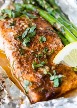 A fillet of salmon sitting on foil with a lemon wedge and asparagus with basil garnish